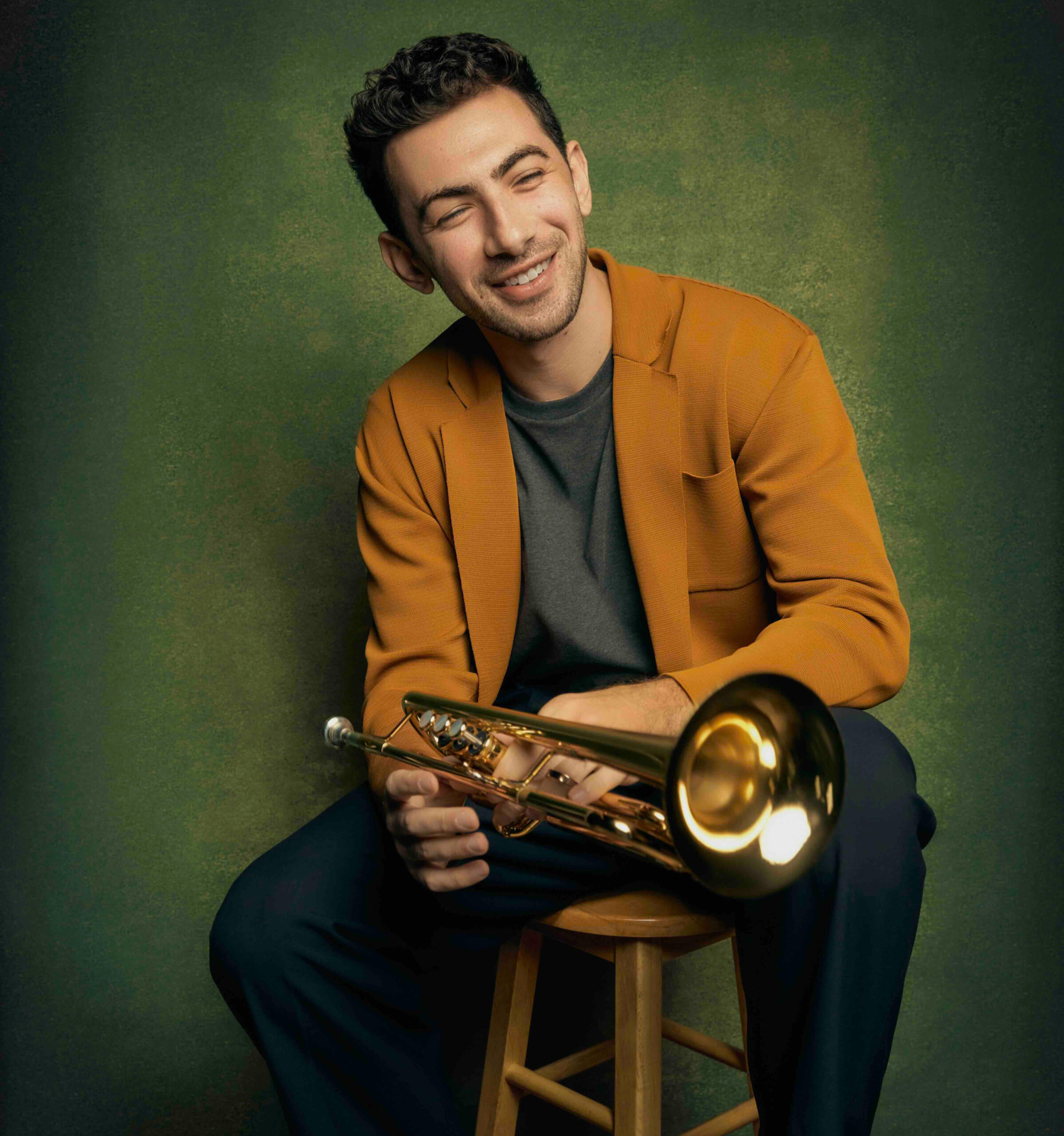 Musician John Dimase sitting wearing autumnal colors and holding his trumpet with a gentle smile. Sitting on a stool. Green backdrop.