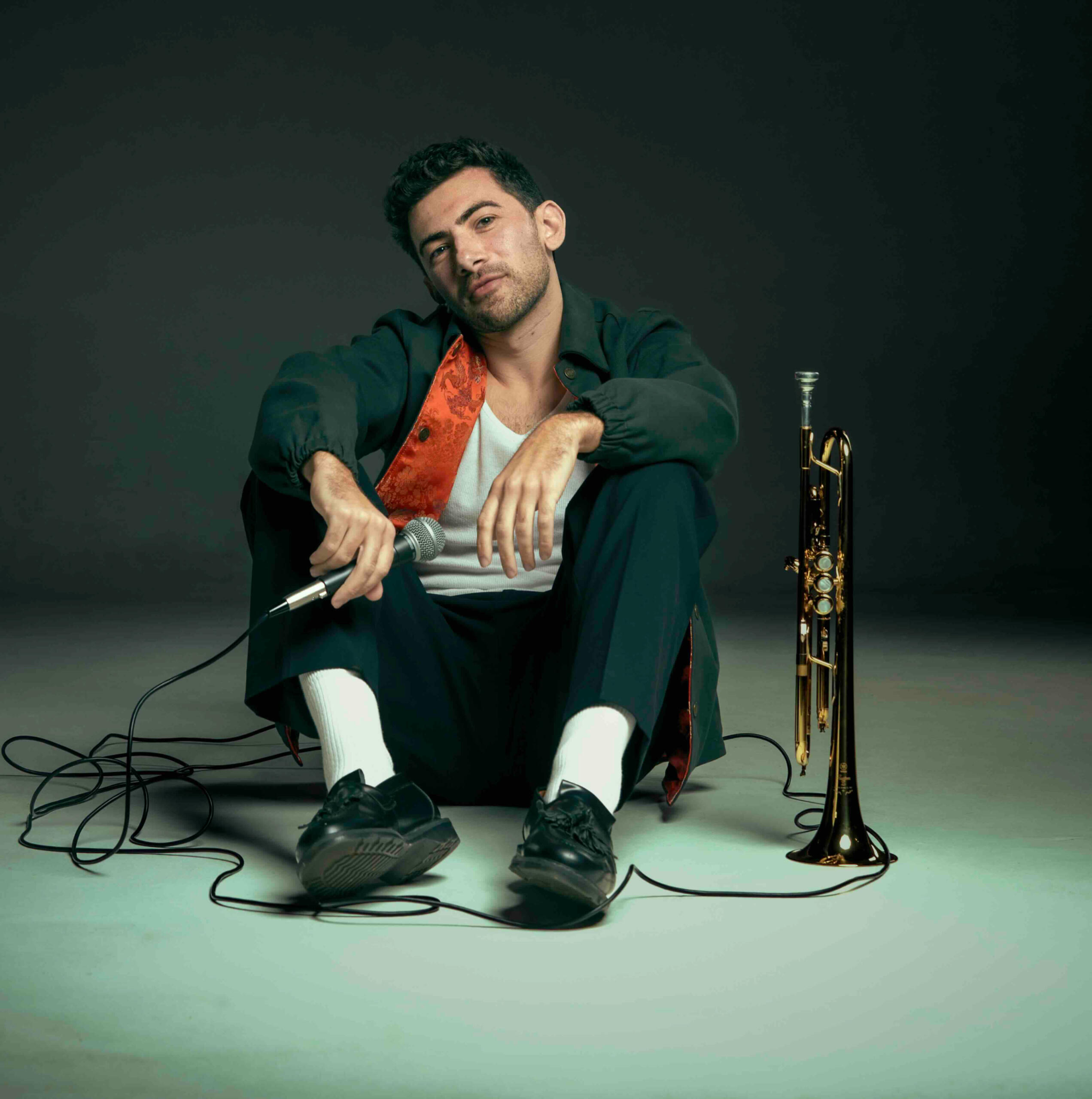 Musician John Dimase sitting down (shot at eye level) wearing a stylish navy jacket with red Chinese styling interior. Holding his microphone and his trumpet is beside him.